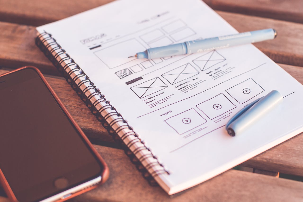 Close-up of a notebook with wireframe sketches and a smartphone on a wooden desk.